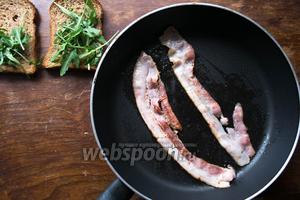 На хлеб (2 кусочка) выложить по небольшому пучку рукколы. Бекон (50 г) обжарить на сильном огне до золотистого цвета.