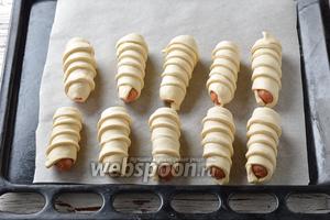 Разрезать длинные сосиски (250 г) вертикально пополам и завернуть каждую половинку в полоску теста, накручивая его по спирали так, чтобы следующий слой слегка заходил на предыдущий. Выложить сосиски в тесте в форму для выпечки с пергаментной бумагой, соблюдая между ними расстояние, так как во время выпечки тесто подрастёт.